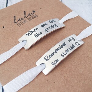 May include: Two silver bracelets with white ribbon ties. The bracelets have engraved messages: "When you feel like quitting" and "Remember why you started".