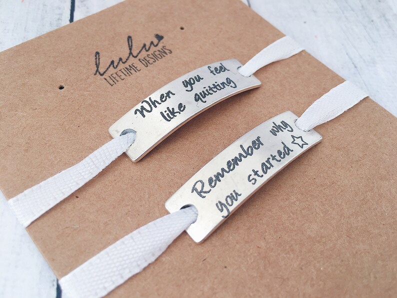 May include: Two silver bracelets with white ribbon ties. The bracelets have engraved messages: "When you feel like quitting" and "Remember why you started".