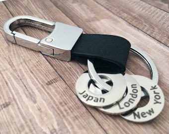 Puede incluir: Un llavero plateado con una correa de cuero negro y tres anillos plateados con las palabras "Japón", "Londres" y "Nueva York" grabadas en ellos.