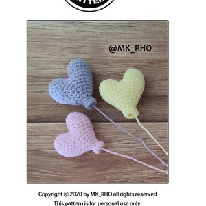 Puede incluir: Tres globos de corazón tejidos a crochet en colores rosa, amarillo y azul. Los globos están atados a cuerdas y se muestran sobre una superficie de madera. La imagen incluye el texto "@MK_RHO" y "Copyright © 2020 by MK_RHO all rights reserved. This pattern is for personal use only. No part of this DESIGN or TEXT may be reproduced, copied, translated or shared without permission."
