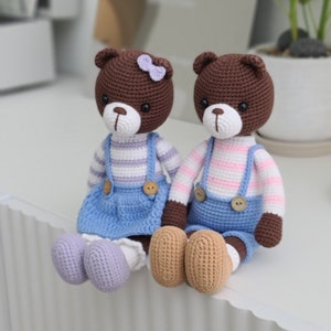 Pode incluir: Dois ursos de peluche de crochet marrons que usam macacões azuis e camisas listradas. Um urso tem um laço roxo na cabeça. Os ursos estão sentados em uma superfície branca.