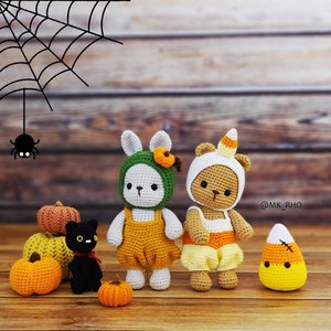 May include: Two crocheted stuffed animals dressed in Halloween costumes. A brown bear wearing a yellow and orange candy corn costume and a white bunny wearing a green and yellow pumpkin costume. There are also several small orange pumpkins and a black cat.