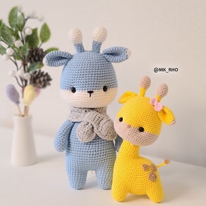 Pode incluir: Dois animais de peluche de crochê, um veado azul com uma echarpe cinza e uma girafa amarela com um laço rosa. O veado tem um rosto branco e olhos pretos. A girafa tem um rosto marrom e olhos pretos. Os animais estão em pé sobre uma superfície branca. @MK_RHO