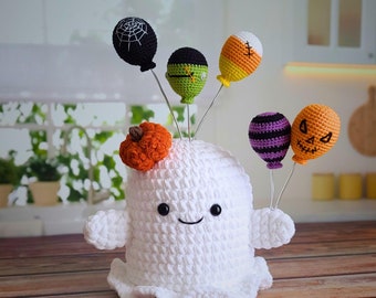Op de afbeelding: Een witte gehaakte geest met een vriendelijk gezicht, die een verscheidenheid aan gehaakte Halloween-decoraties vasthoudt, waaronder een pompoen, een spinnenweb, een maïs, een geest en een paarse ballon.