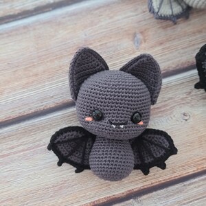 Puede incluir: Un juguete de peluche de murci&eacute;lago tejido a crochet con alas negras y una cara sonriente linda. El murci&eacute;lago es gris con ojos negros y un rubor rosado en las mejillas.