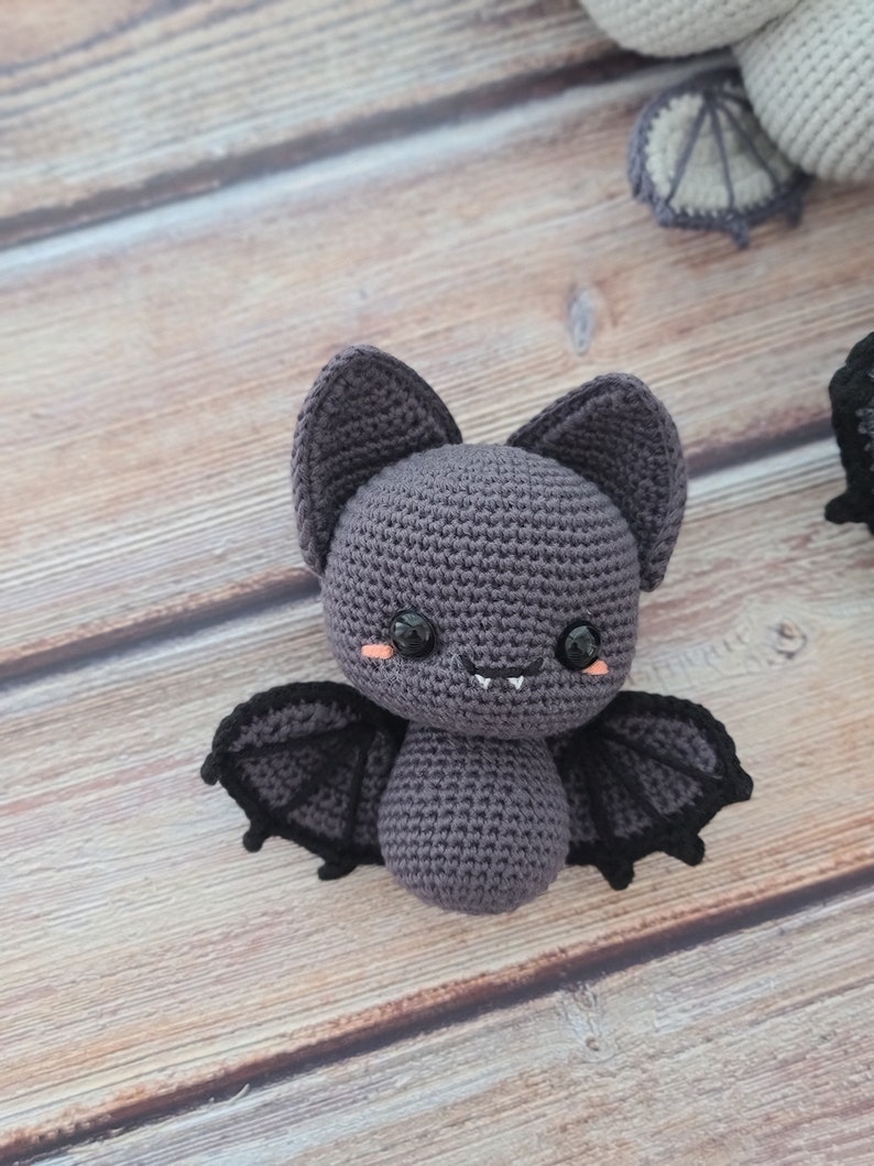 Puede incluir: Un juguete de peluche de murci&eacute;lago tejido a crochet con alas negras y una cara sonriente linda. El murci&eacute;lago es gris con ojos negros y un rubor rosado en las mejillas.