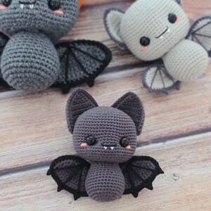Puede incluir: Tres murci&eacute;lagos de peluche tejidos a crochet con ojos negros, mejillas rosadas y dientes blancos. Los murci&eacute;lagos son grises, gris claro y gris oscuro. Los murci&eacute;lagos est&aacute;n sentados sobre una superficie de madera.