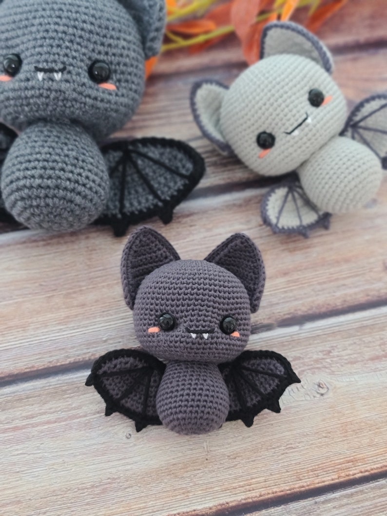 Puede incluir: Tres murci&eacute;lagos de peluche tejidos a crochet con ojos negros, mejillas rosadas y dientes blancos. Los murci&eacute;lagos son grises, gris claro y gris oscuro. Los murci&eacute;lagos est&aacute;n sentados sobre una superficie de madera.