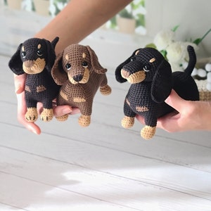 May include: Three crocheted dachshund dog toys in black, brown, and black and brown. The toys are all sitting on a white wooden surface.