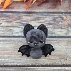 Puede incluir: Un murci&eacute;lago de crochet gris con alas negras y ojos rosas. El murci&eacute;lago est&aacute; sentado sobre una superficie de madera.