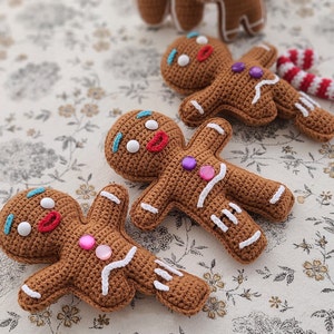 May include: Three crocheted gingerbread people with white icing details and colourful buttons. The gingerbread people are lying on a white background with a floral pattern.