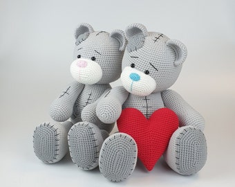 May include: Two gray crocheted teddy bears holding a red heart. The bears are sitting side by side and looking at each other.