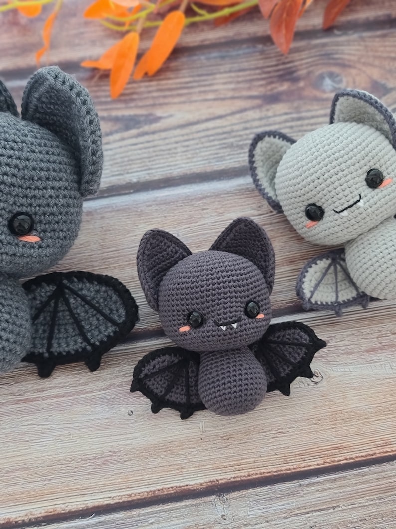 Puede incluir: Tres murci&eacute;lagos de crochet de diferentes tama&ntilde;os, todos con ojos negros y mejillas rosadas. El murci&eacute;lago m&aacute;s grande es gris con alas negras, el murci&eacute;lago del medio es gris oscuro con alas negras y el murci&eacute;lago m&aacute;s peque&ntilde;o es gris claro con alas blancas.
