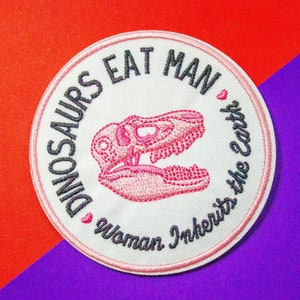 May include: White embroidered patch with the text "Dinosaurs Eat Man ♥ Woman Inherits the Earth" and a pink dinosaur skull graphic.