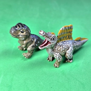 以下が含まれることがあります： 2体の小さな恐竜のフィギュア。1体は暗い縞模様のある茶色で質感のあるティラノサウルス・レックス。もう1体は、目立つ黄色と黒の背びれ、長い鼻先、赤い口の中が見える開いた口、そして灰色の斑点模様の皮膚を持つスピノサウルスです。