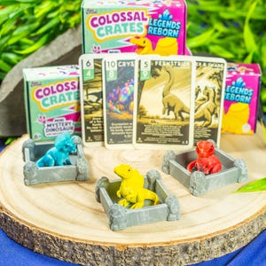 Może przedstawiać: Plaster drewna prezentuje miniaturowe figurki dinozaurów w szarych skrzyniach, z kartami kolekcjonerskimi i pudełkami z napisem "Colossal Crates Legends Reborn". Dinozaury są niebieskie, żółte i czerwone. Zielone liście i niebieskie tło dopełniają scenę.