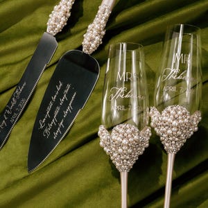 May include: A wedding cake knife and server set with two champagne flutes. The handles of the knife and server are adorned with pearls and rhinestones. The champagne flutes have pearl accents and are engraved with names and a date.