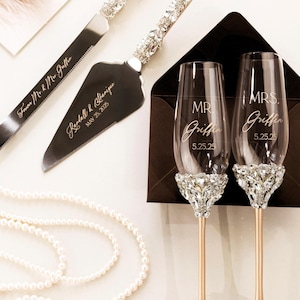 Może przedstawiać: Zestaw noża i serwera do ciasta weselnego z kryształowymi akcentami oraz wygrawerowanymi imionami i datą. Dwa kieliszki do szampana z kryształowymi podstawami i złotymi nóżkami, z grawerem "MR" i "MRS" oraz datą. Perłowy naszyjnik i artykuły papiernicze.