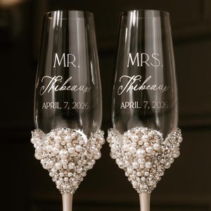 May include: Two champagne flutes with pearl and crystal embellishments at the base. The flutes are engraved with "MR. Thibeau" and "MRS. Thibeau" and the date "APRIL 7, 2026". The stems are a light cream color.