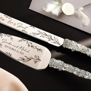 May include: A silver cake cutting set with a floral design and rhinestones. The knife has the text "Love is Sweet" and the server has the text "Sandra and Michael September 23, 2023".