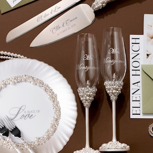 May include: Wedding cake knife and server set with pearl and rhinestone accents. Includes two champagne flutes, a plate, and forks. The plate and flutes have pearl embellishments. The cake server is engraved with "A Slice of Love" and a name and date.