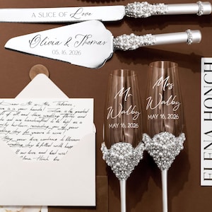 May include: A wedding-themed still life featuring a cake knife and server set, two champagne flutes, and a handwritten letter. The knife and server are silver with crystal accents. The flutes are adorned with crystals and personalized with names and a date.