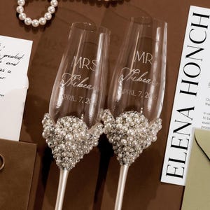 May include: Two champagne flutes with pearl embellishments. The flutes are engraved with "MRS" and "MR", names, and the date "APRIL 7, 2016". The glasses sit on a brown surface with wedding-related items.