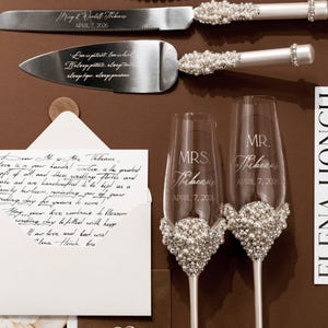 May include: A wedding cake knife and server set with pearl and rhinestone accents. Two champagne flutes with pearl embellishments and the text "MRS. Tribeau" and "MR. Tribeau" are also present. A handwritten letter and wedding rings are included.