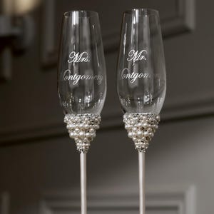May include: Two champagne flutes with pearl and rhinestone embellishments. The glasses are engraved with "Mrs. Montgomery" and "Mr. Montgomery" in elegant script, perfect for a wedding or anniversary celebration.