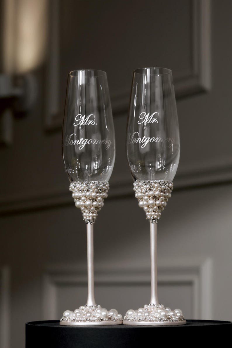 May include: Two champagne flutes with pearl and rhinestone embellishments. The glasses are engraved with "Mrs. Montgomery" and "Mr. Montgomery" in elegant script, perfect for a wedding or anniversary celebration.