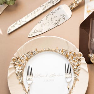 Pode incluir: Um prato de cerâmica branco com uma borda de strass dourados e transparentes. Dois garfos de prata com as palavras "MR." e "MRS." gravadas nas alças estão colocados no prato. O prato tem as palavras "Arian & Albertine 21.01.2014" escritas em itálico no centro.