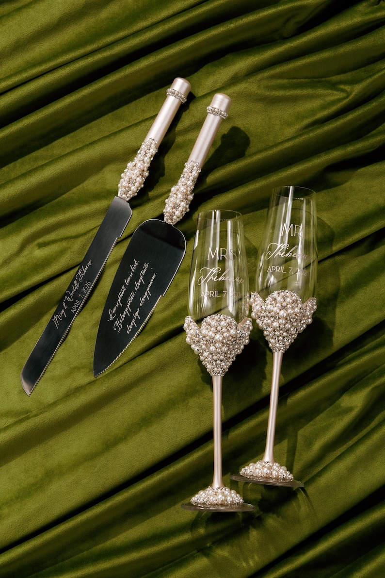 May include: A wedding cake knife and server set with two champagne flutes. The handles of the knife and server are adorned with pearls and rhinestones. The champagne flutes have pearl accents and are engraved with names and a date.