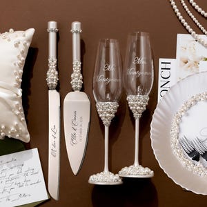 May include: Wedding accessories including a white satin ring pillow, cake knife and server, champagne flutes, and a decorative plate. The knife and server have pearl accents. The flutes are engraved with a name. The plate has a pearl border.