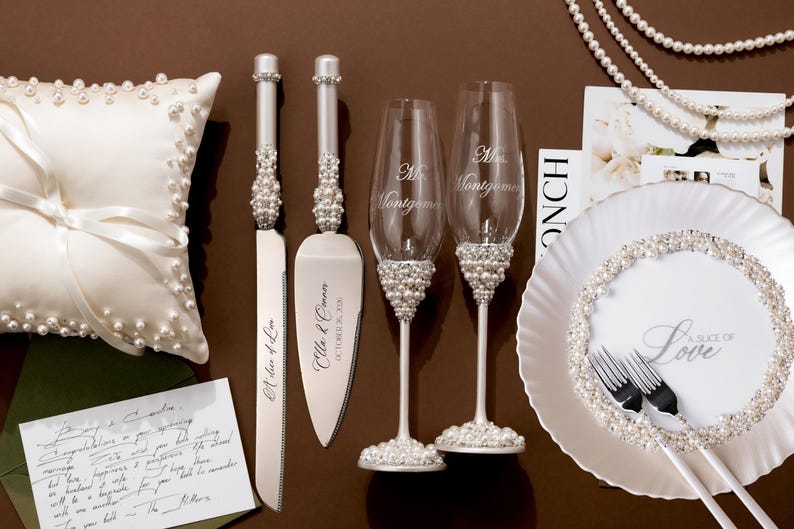 May include: Wedding accessories including a white satin ring pillow, cake knife and server, champagne flutes, and a decorative plate. The knife and server have pearl accents. The flutes are engraved with a name. The plate has a pearl border.