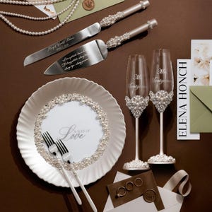 May include: Wedding cake knife and server set with pearl accents, alongside two champagne flutes, a plate, and forks. The plate features the text "A Slice of Love". The flutes are decorated with pearls and the names "Mr. & Mrs. Montgomery".