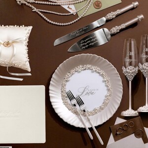May include: Wedding-themed items arranged on a brown surface. Includes a ring pillow, cake server and knife set, champagne flutes, a plate with forks, a guest book, and wedding rings. The plate has the word "Love" on it.