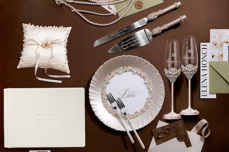 May include: Wedding-themed items arranged on a brown surface. Includes a ring pillow, cake server and knife set, champagne flutes, a plate with forks, a guest book, and wedding rings. The plate has the word "Love" on it.