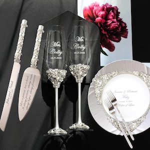 May include: A wedding set comprising a cake knife and server, two champagne flutes, and a decorative plate with forks. The knife and server have crystal-studded handles. The flutes and plate are adorned with crystal accents. The set is personalized with names and a date.