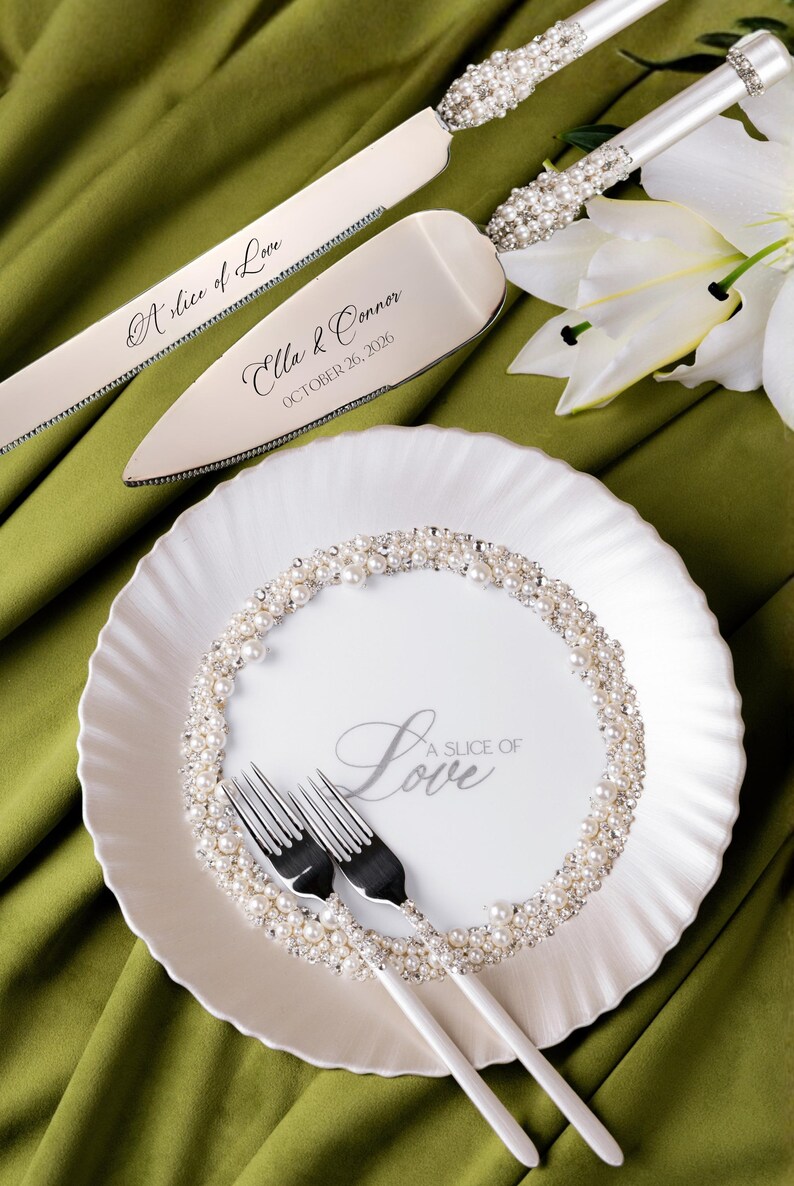 May include: A wedding cake serving set and plate with forks. The silver cake knife and server are adorned with pearl accents and the words "A slice of Love" and names with a date. The white plate has a pearl border and the words "A Slice of Love".