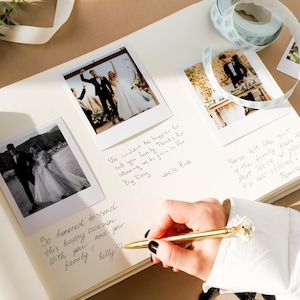May include: An open wedding guest book with polaroid photos and handwritten notes. A gold pen is held over the page, ready to write. Decorative ribbon and sprigs of greenery are in the background.