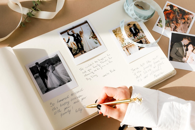 May include: An open wedding guest book with polaroid photos and handwritten notes. A gold pen is held over the page, ready to write. Decorative ribbon and sprigs of greenery are in the background.