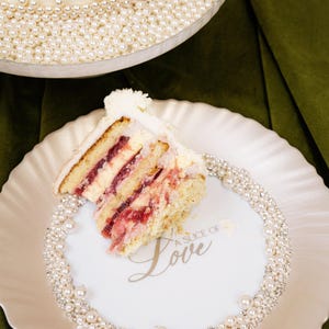 May include: A slice of layered cake with white frosting and red filling on a white plate. The plate is decorated with pearls and the words "A Slice of Love". A larger cake with pearl accents is in the background.
