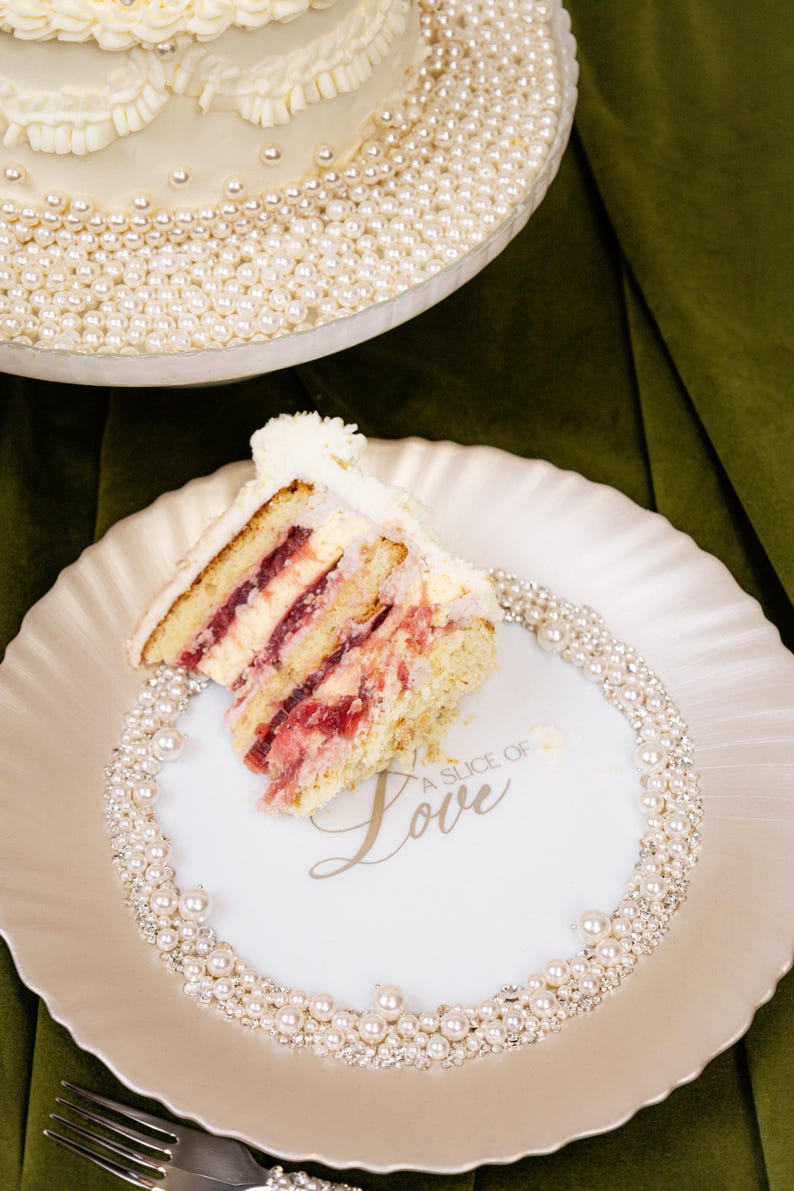 May include: A slice of layered cake with white frosting and red filling on a white plate. The plate is decorated with pearls and the words "A Slice of Love". A larger cake with pearl accents is in the background.