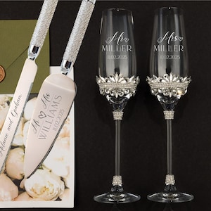 Puede incluir: Un juego de cuchillo para cortar pastel de boda con dos copas de champán. El cuchillo para pastel tiene un mango plateado con diamantes de imitación y las palabras "Mr & Mrs WILLIAMS 11.02.2025" grabadas en la hoja. Las copas de champán tienen bordes plateados con diamantes de imitación y las palabras "Mr MILLER 11.02.2025" y "Mrs MILLER 11.02.2025" grabadas en los vasos.