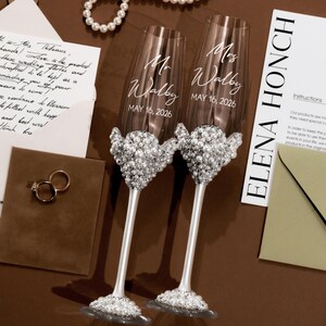 May include: Two champagne flutes with pearl embellishments. Each glass is engraved with "Mr Walby" and "Mrs Walby" and the date "MAY 16, 2026". The flutes are on a brown surface, with a wedding invitation and rings.