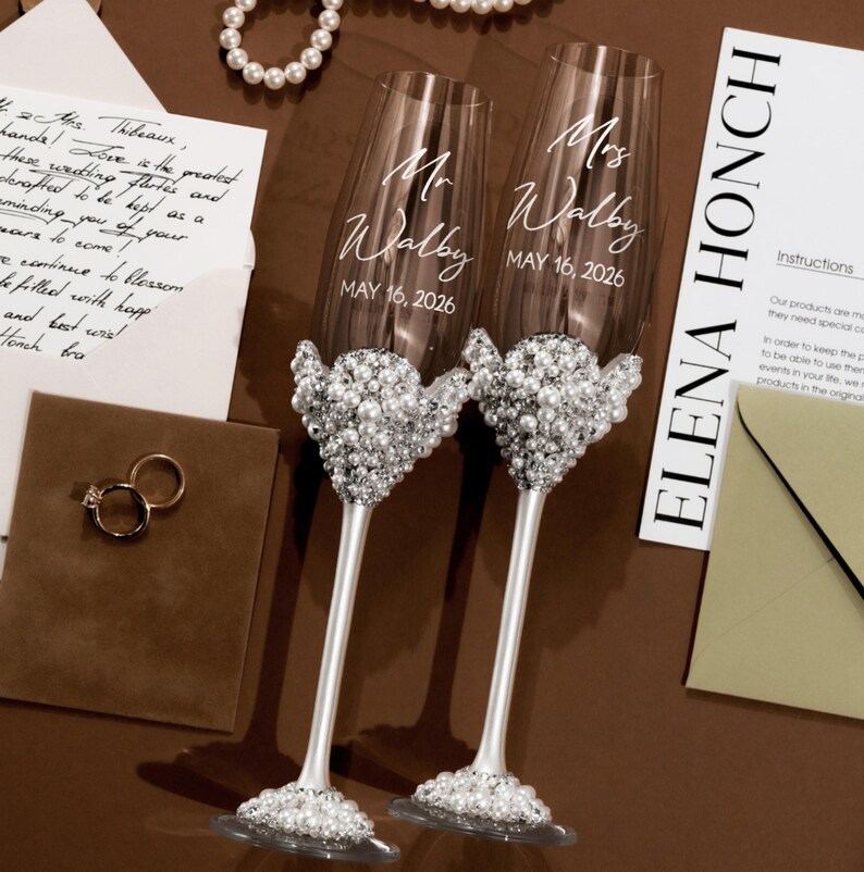 May include: Two champagne flutes with pearl embellishments. Each glass is engraved with "Mr Walby" and "Mrs Walby" and the date "MAY 16, 2026". The flutes are on a brown surface, with a wedding invitation and rings.