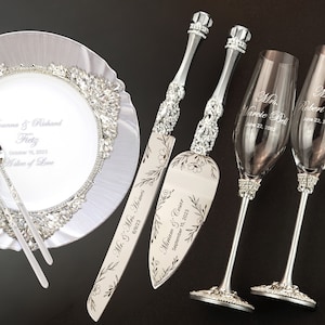 May include: A wedding cake cutting set with two silver knives and two champagne flutes. The knives have floral designs and are engraved with the names "Mr. & Mrs. Homes" and "Witam & Criar" along with their wedding dates. The champagne flutes are also engraved with the names "Mr. Robert Bell" and "Mrs. Warete Bell" and their wedding date.