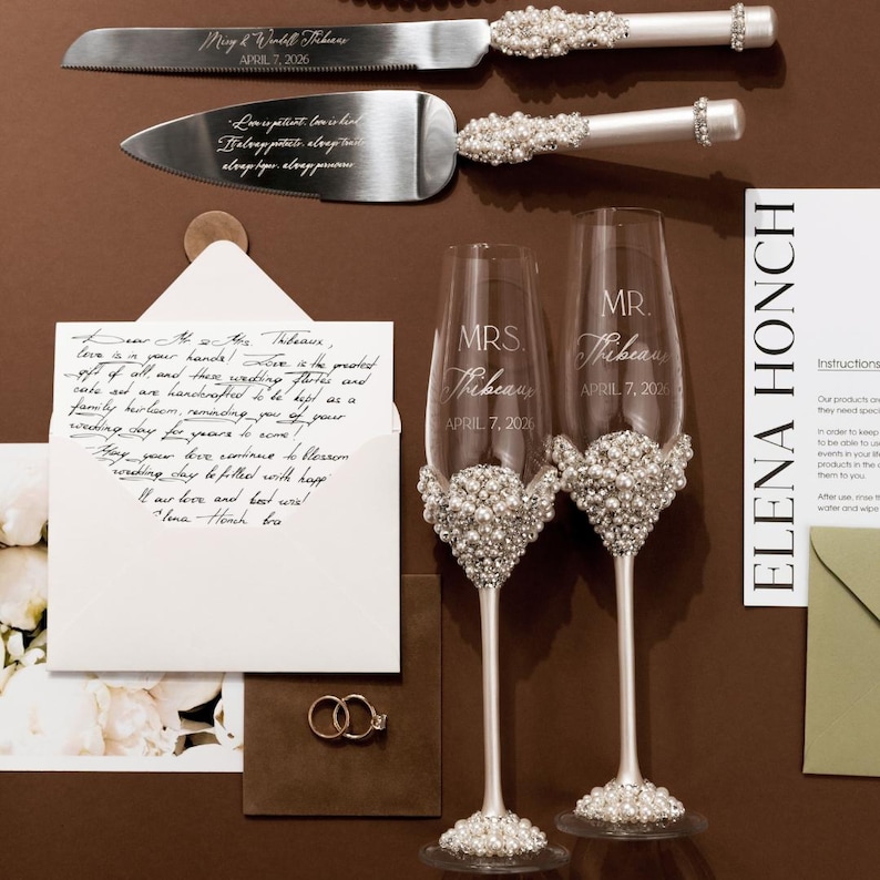 May include: A wedding cake knife and server set with pearl and rhinestone accents. Two champagne flutes with pearl embellishments and the text "MRS. Tribeau" and "MR. Tribeau" are also present. A handwritten letter and wedding rings are included.