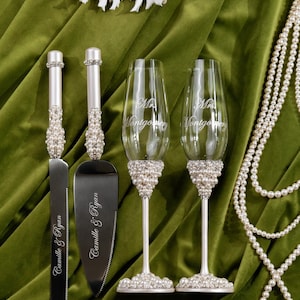 May include: Wedding cake knife and server set with pearl and rhinestone accents. Two champagne flutes with pearl bases and the text "Mr. Montgomery" are also shown. A string of pearls and a seashell are also in the image.
