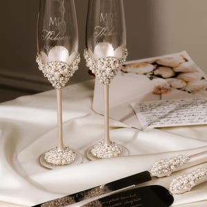 May include: Elegant champagne flutes and cake serving set with pearl embellishments. The flutes are engraved with "MR" and "MRS" and a last name. The cake server and knife have pearl accents. Wedding theme.